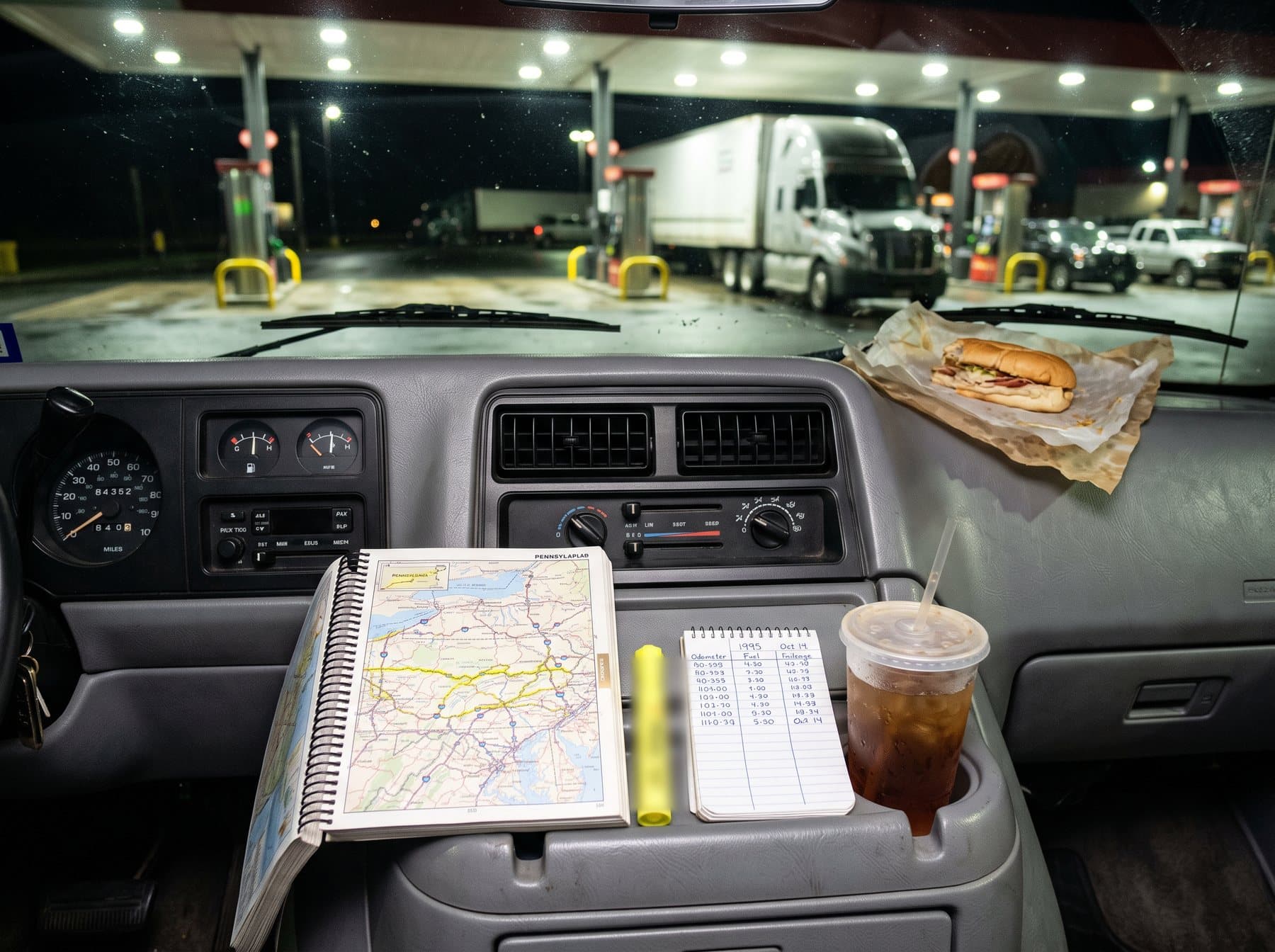 A road atlas, a yellow highlighter and a small fuel notebook on the dashboard of a late-1980s van